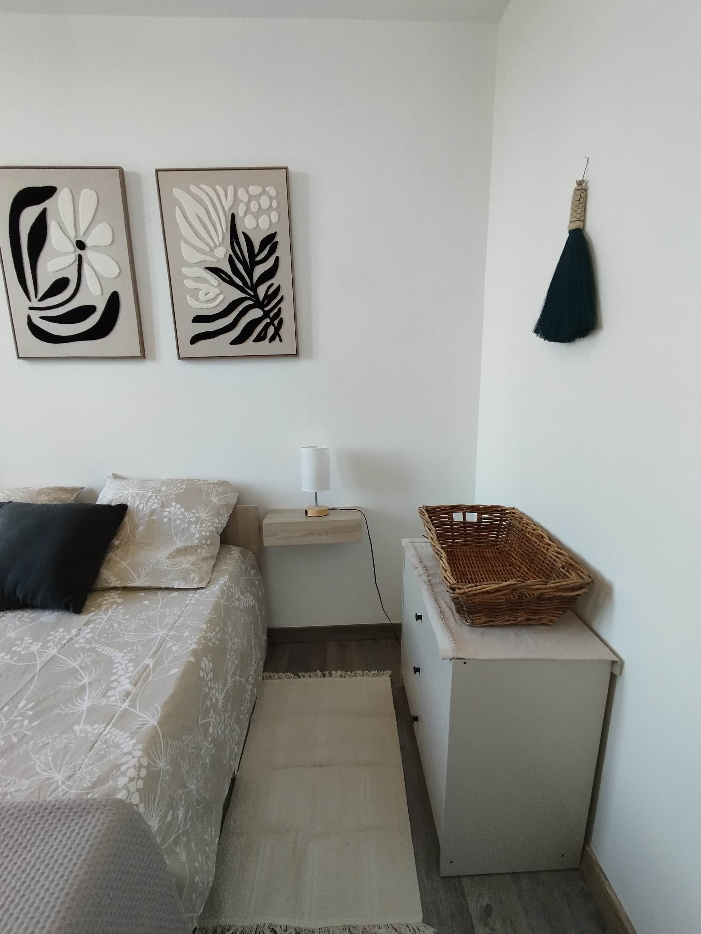 Une chambre à coucher comprenant un lit, une table de chevet, une lampe, une frange décorative fixée au mur et un panier en osier.