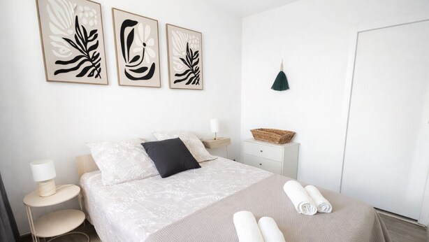 Una camera da letto ordinata con un letto, cuscini, un comodino, una lampada, un cesto a parete e una porta.