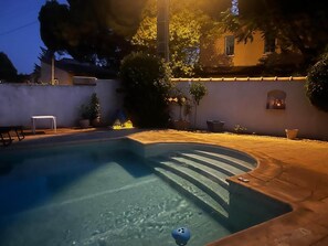 A swimming pool with a curved edge, surrounded by a paved patio, potted plants, and a building with a window. - La Bastide des Gayants : garden, swimming pool, wifi (Le Luc)