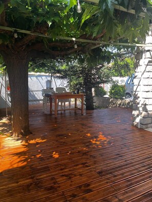 A wooden deck with a table and chairs, surrounded by trees and a stone wall. - La Bastide des Gayants : garden, swimming pool, wifi (Le Luc)