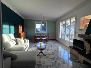 A living room with a sofa, a dining table with chairs, a fireplace, and large windows. - La Bastide des Gayants : garden, swimming pool, wifi (Le Luc)