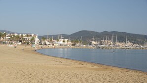 Una playa con un puerto deportivo, edificios y montañas al fondo.