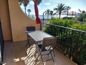 A balcony with a table and chairs, overlooking a beach with palm trees and flags. - Waterfront studio: sea view, air conditioning, wifi (Cogolin)