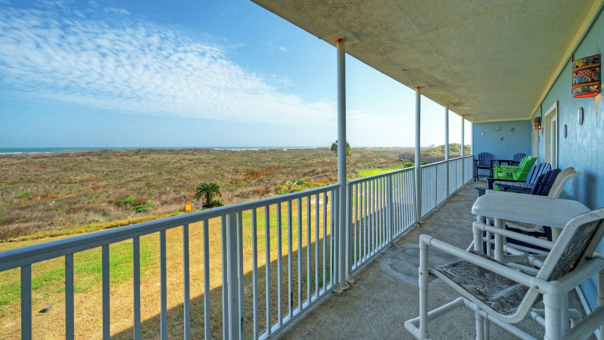 A covered balcony with a view of the beach, white railings, and outdoor furniture.