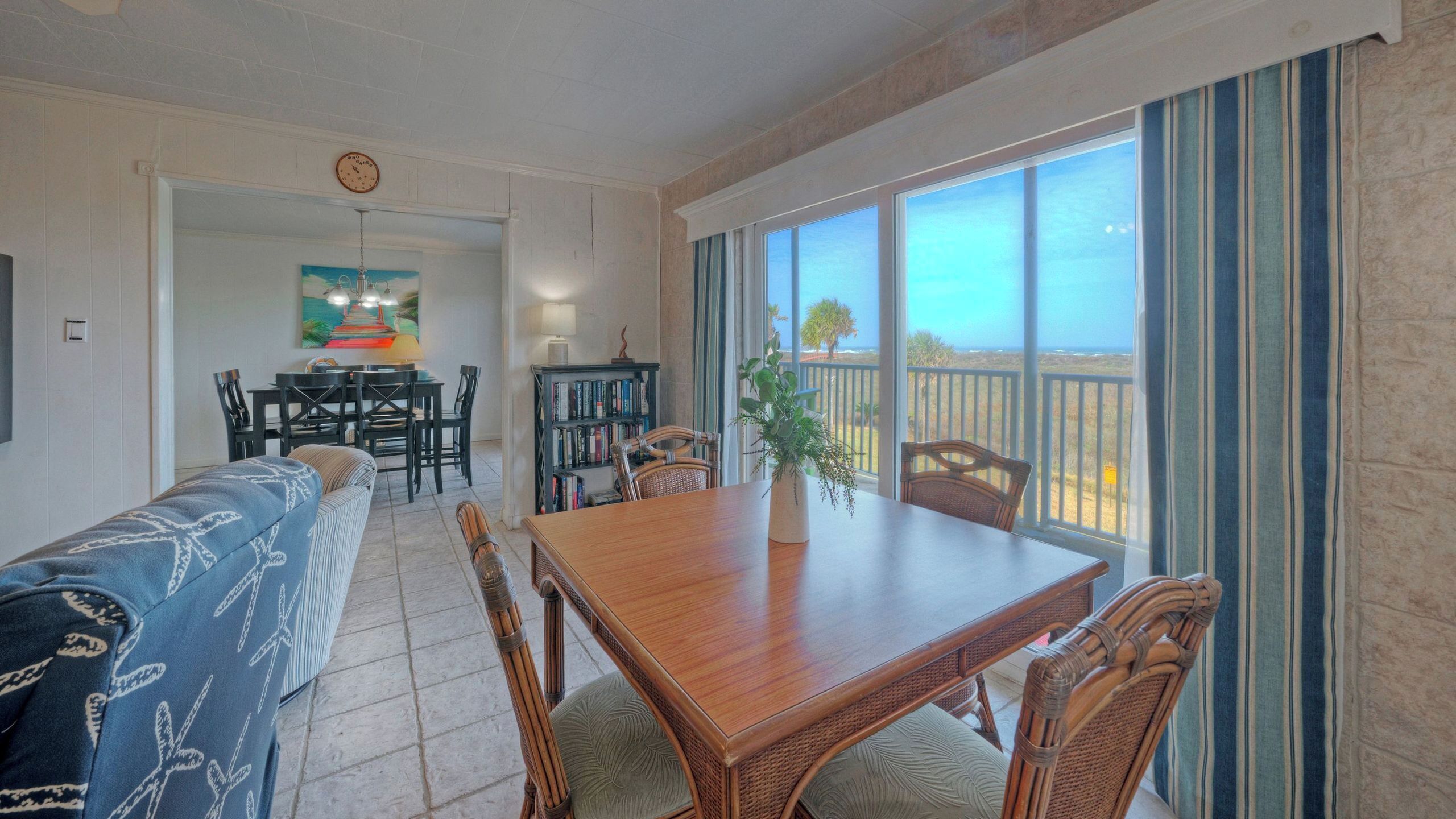 A dining area with a wooden table and chairs, a sofa, a bookshelf, and a view of the outdoors.