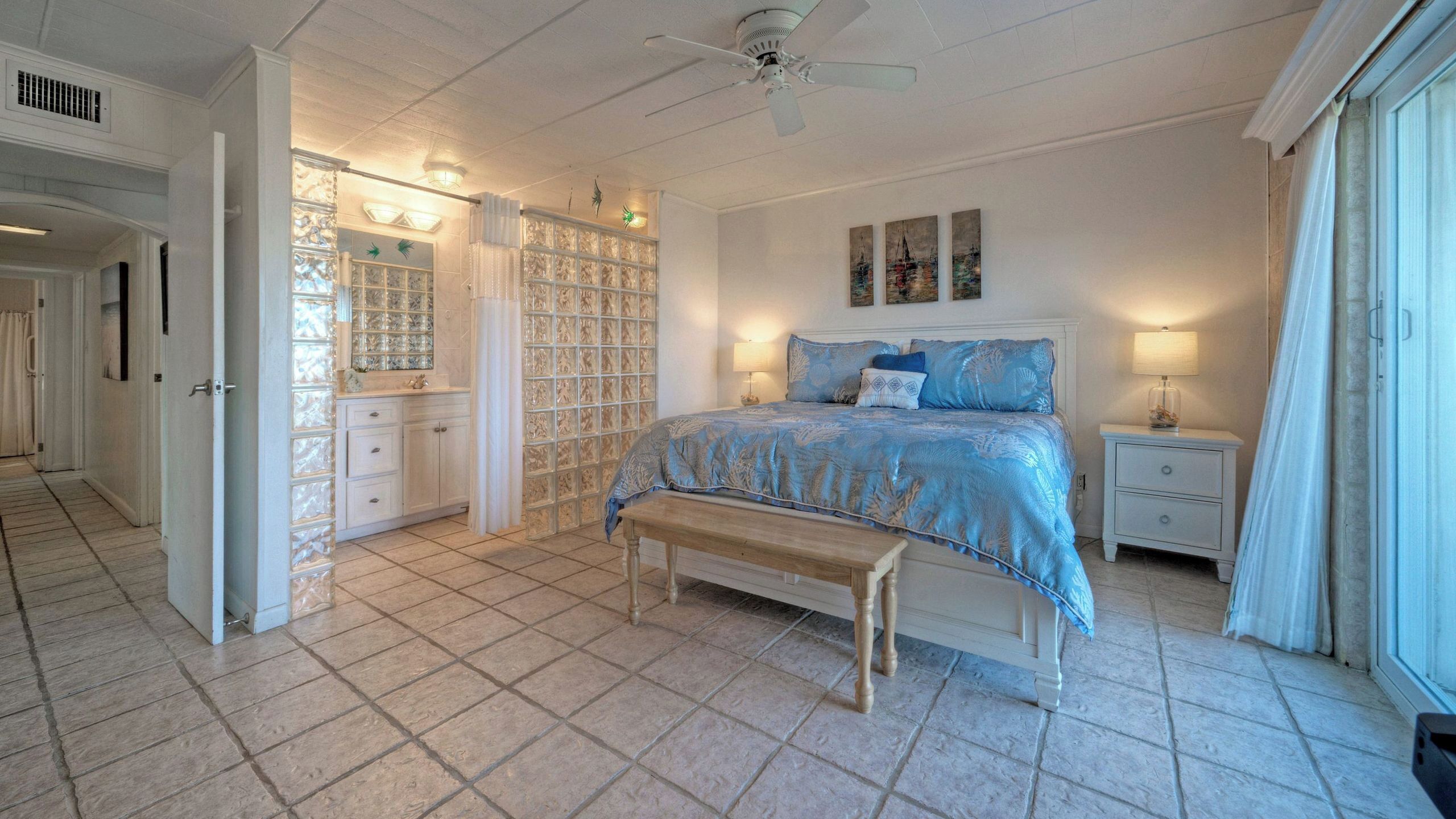 Une chambre à coucher comprenant un lit, des tables de chevet, un banc, un ventilateur de plafond et une vue sur l’extérieur.