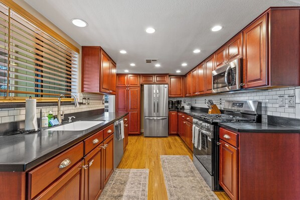 A modern kitchen with wooden cabinets, stainless steel appliances, a black countertop, and a window with blinds. - NEW! Casa de Cameron Games/Grill/Lake! (Flagstaff)