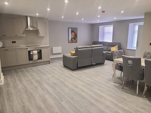 Een moderne woonkamer met een grijze bank, een eettafel met stoelen, een keuken met kasten en een fornuis, en een schilderij aan de muur.