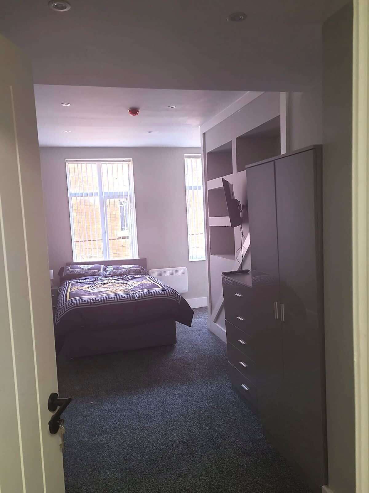 Un dormitorio con una cama, un aparador, un televisor montado en la pared y una ventana con persianas.