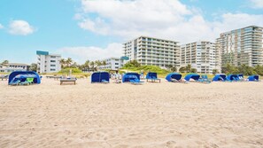 A beach with multiple blue tents and lounge chairs, surrounded by high-rise buildings. - Pompano Beach | Coastal Ocean Retreat | Pool + Beach (Pompano Beach)