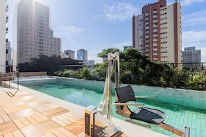 Ein Pool auf dem Dach mit einer Holzterrasse, einer Liege und Blick auf umliegende Hochhäuser.