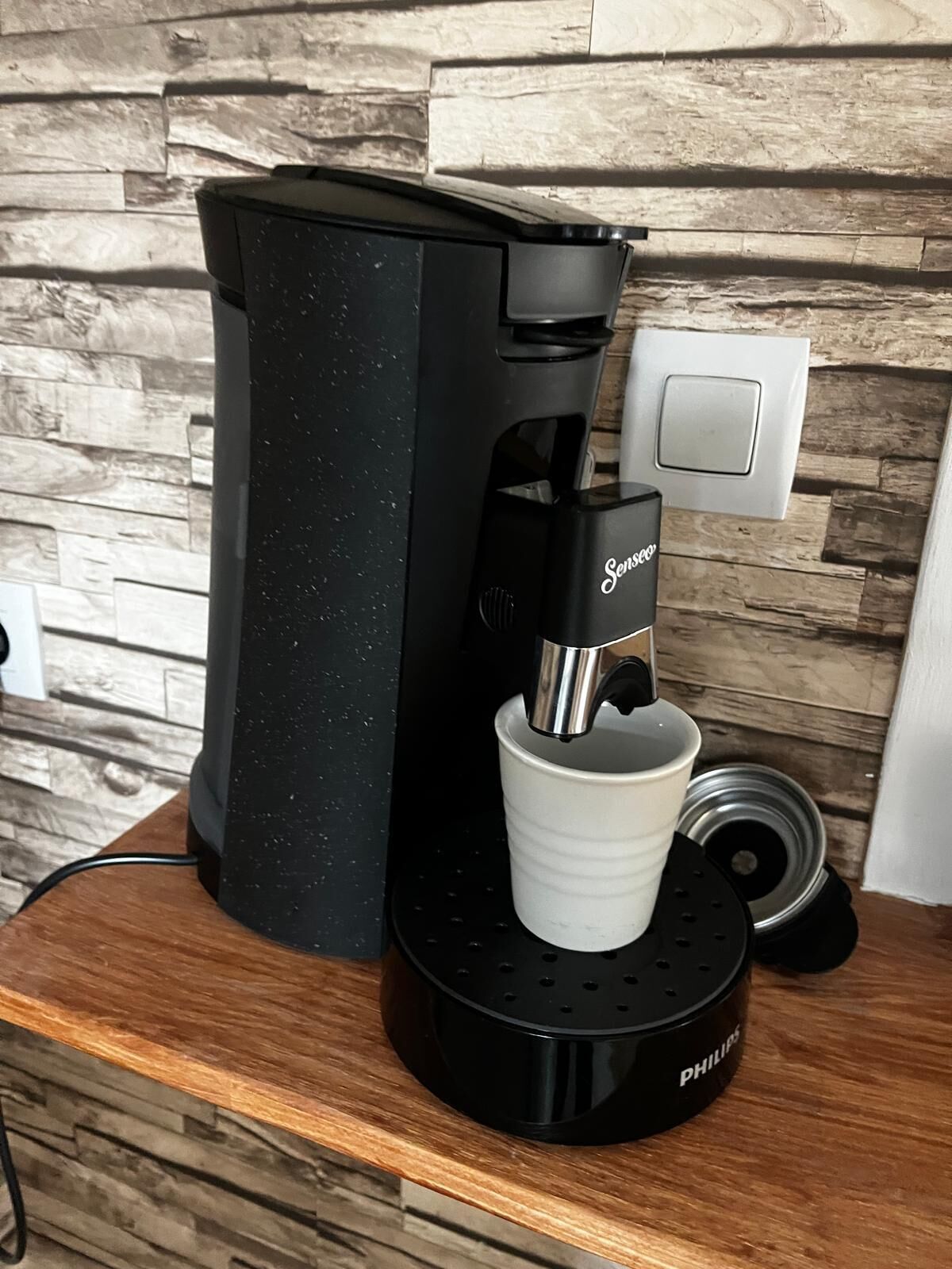 A coffee maker with a white cup holder, a power cord, and a wall socket.
