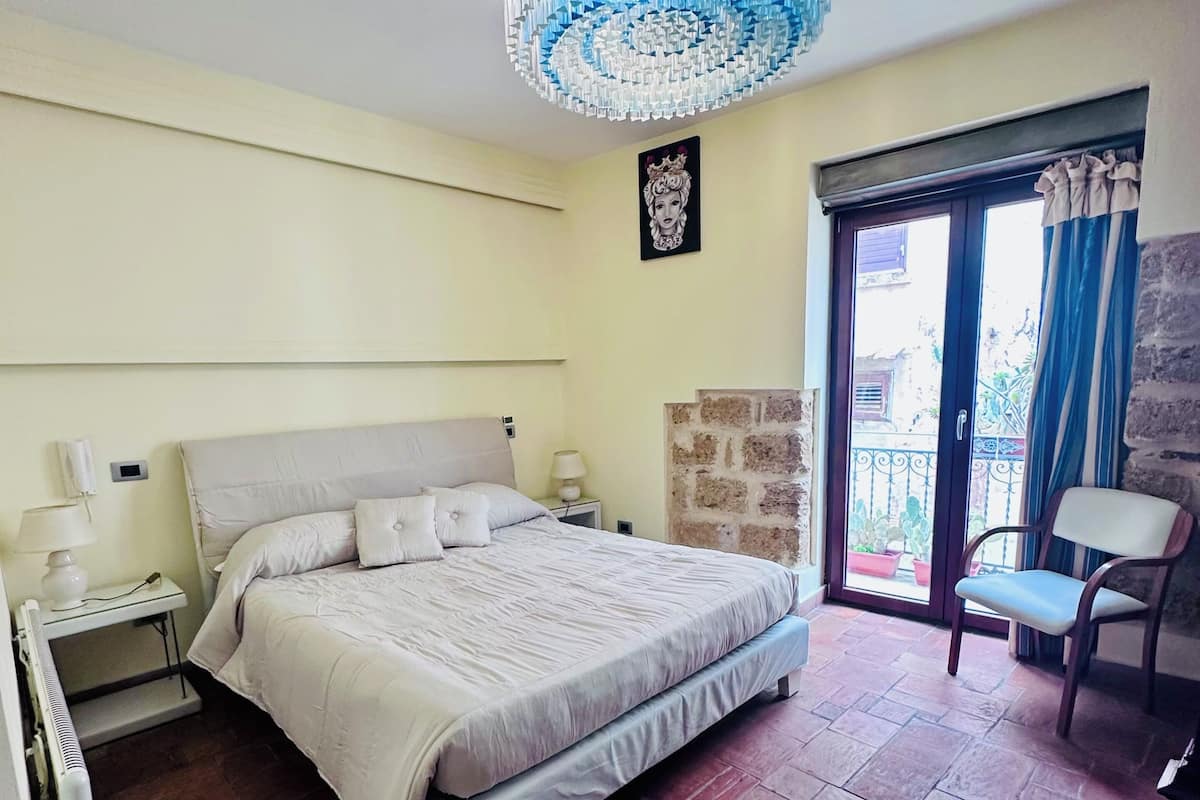 Un dormitorio con cama, mesitas de noche, una araña y una silla, con vistas a través de un ventanal.