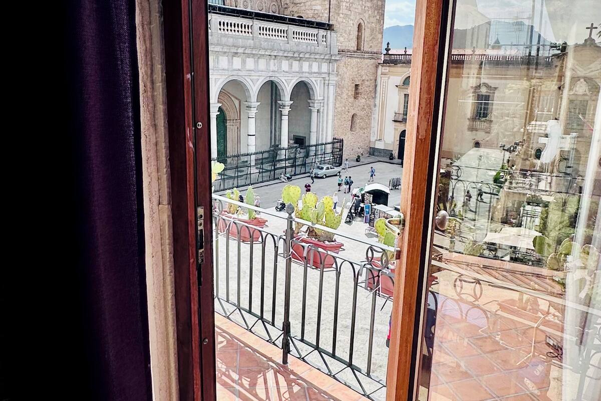 Vista a través de una ventana que muestra un edificio con patio interior, una torre alta y un área cercada con plantas de cactus.