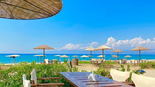 Ein Essbereich am Strand mit Holztischen und -stühlen, umgeben von Reetdachschirmen und Blick auf den Ozean.