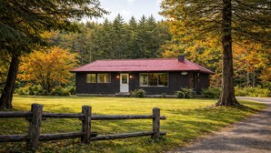 A rustic cabin with a red roof, surrounded by a wooden fence, set in a forest with autumn foliage.