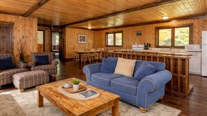 A cozy cabin interior with a wooden dining area, a blue sofa, plaid armchairs, and a kitchen with a microwave.