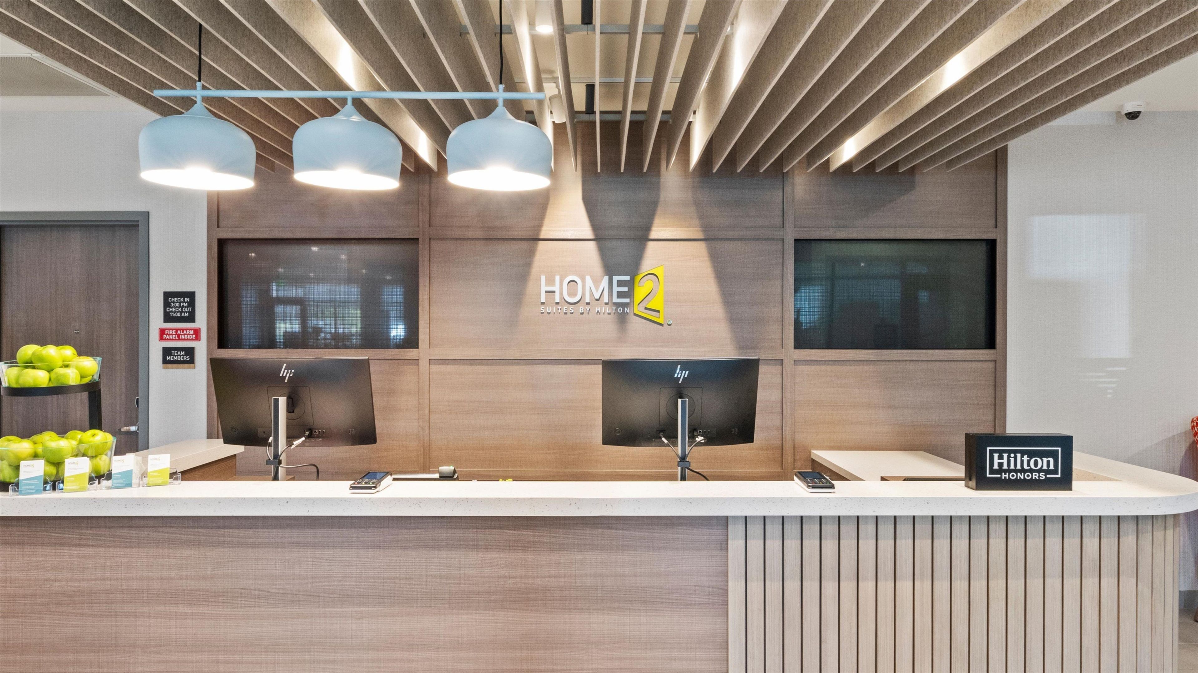 A hotel reception area with a modern design, including a reception desk, multiple monitors, and a sign for 'HOME 2 Suites by Hilton'.