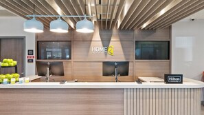 Un'area reception di un hotel dal design moderno, con una scrivania della reception, diversi monitor e un cartello con la scritta 'HOME 2 Suites by Hilton'.