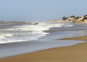 A beach with waves, sandy shore, and buildings in the background.