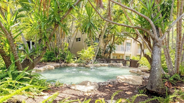 A natural pool surrounded by lush greenery and trees, with a building in the background.