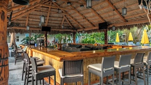 Un área de bar con mostrador de madera y sillas de mimbre, rodeada de plantas tropicales y un techo de paja.