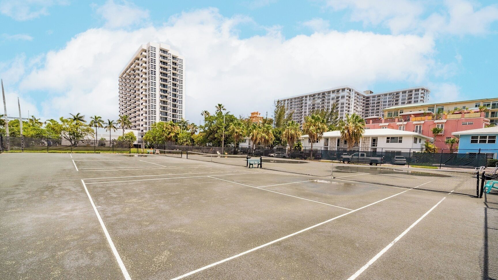An outdoor tennis court with a net, surrounded by buildings and palm trees.