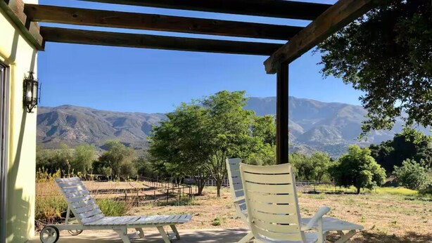 A covered outdoor seating area with two white lounge chairs and a table, overlooking a vineyard and mountains. - Ojai’s Sage Ranch Guest Villa (Ojai)