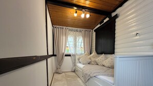 Ein schmaler Raum mit einem Bett, einem Fenster mit Vorhängen, einem Heizkörper und einer Decke mit Holzbalken und Lampen.