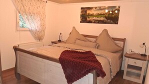 Ein Schlafzimmer mit Bett, Nachttisch, Lampe, einem Fenster mit Vorhängen und einem Wandbild.