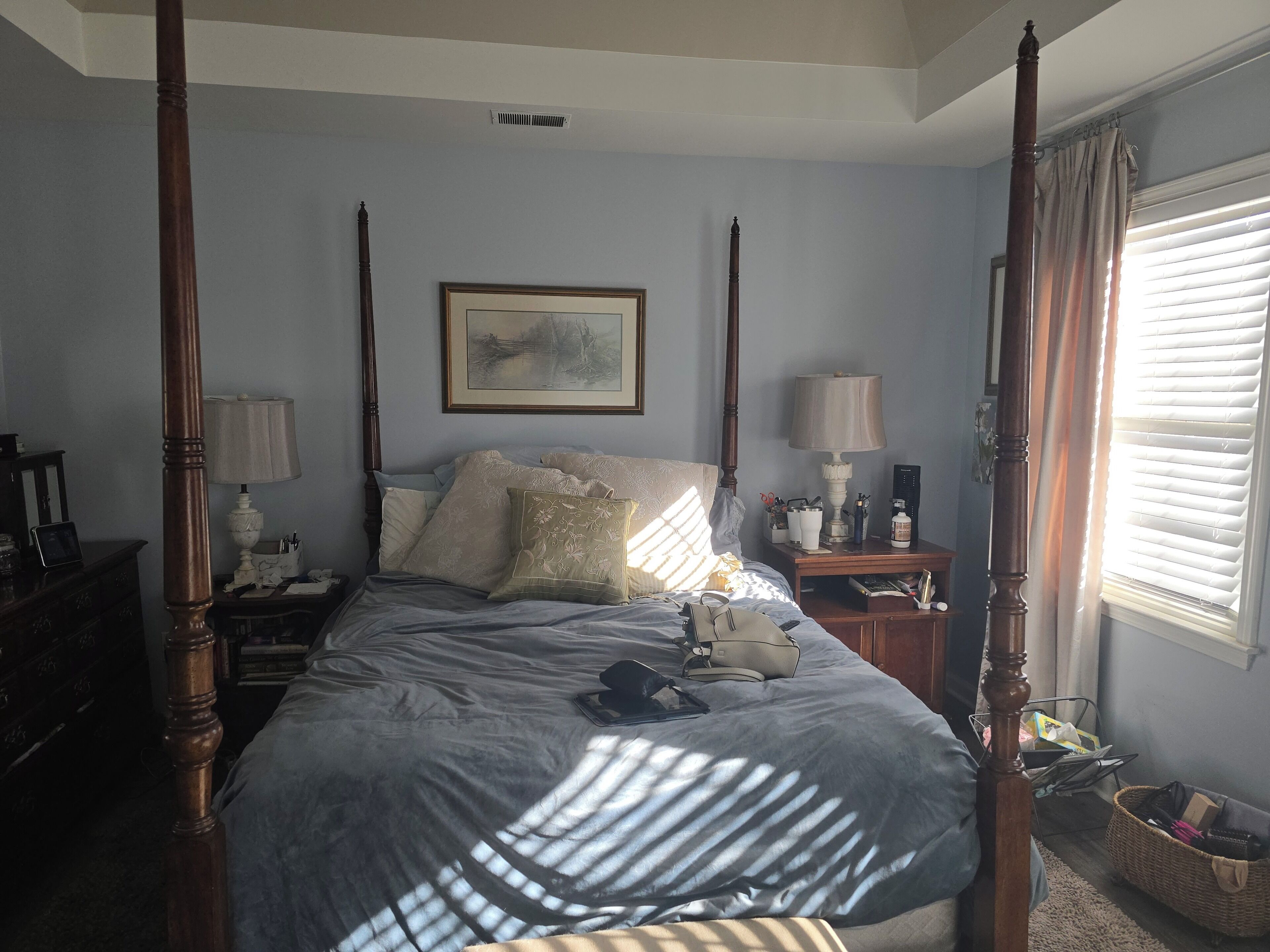 A four-poster bed with a headboard, a bedside table with a lamp, a framed picture on the wall, and a window with blinds.