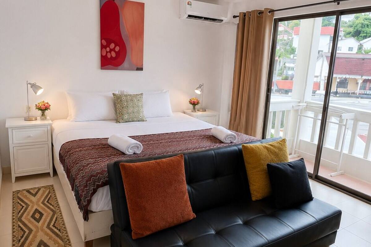 Habitación de hotel con cama, sofá negro, ventana con vistas, mesita de noche y aire acondicionado instalado en la pared.