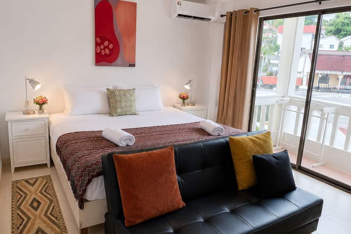 A hotel room with a bed, a black sofa, a window with a view, a nightstand, and a wall-mounted air conditioner.