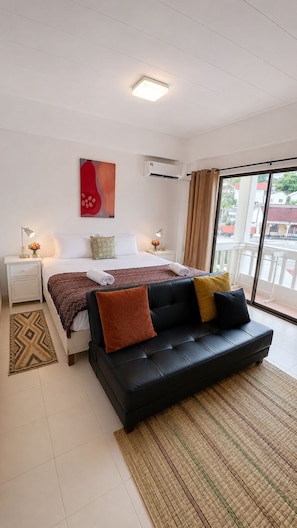Habitación de hotel con cama, sofá negro, ventana con vistas, mesita de noche y aire acondicionado instalado en la pared.