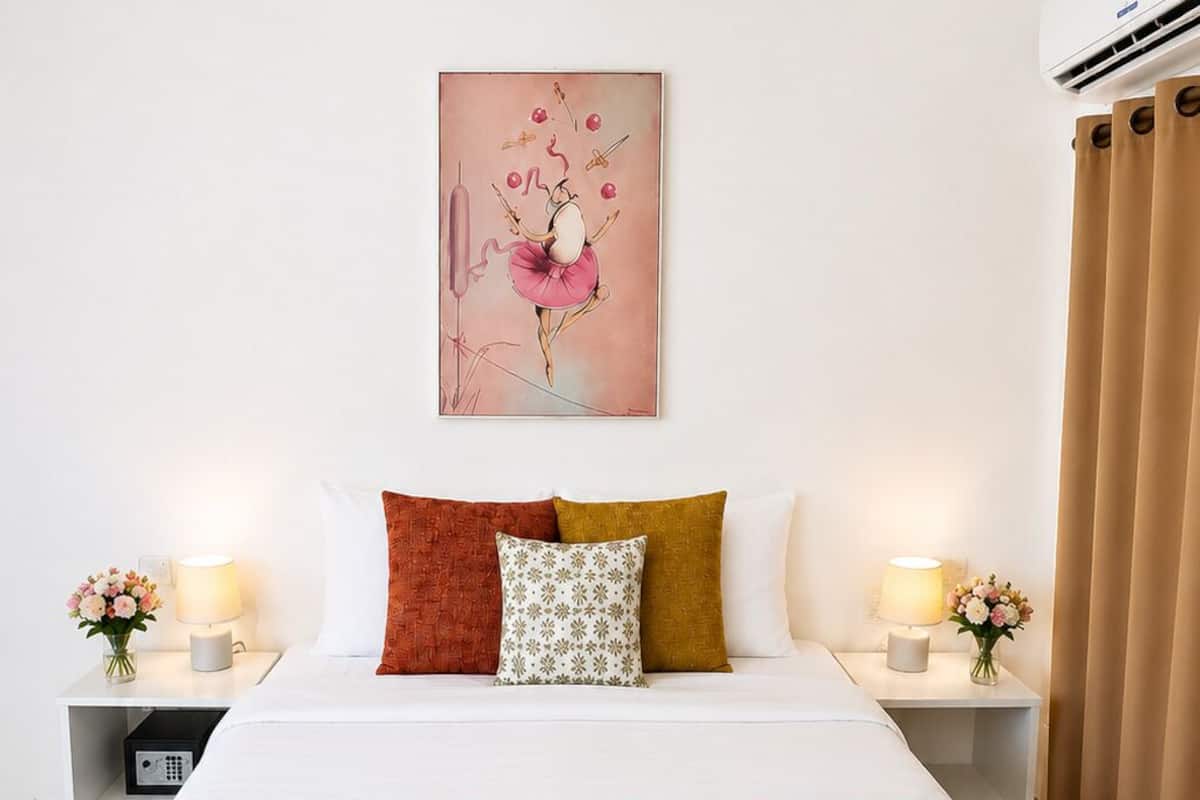 A hotel room with a bed, pillows, a painting, bedside lamps, a nightstand, and a window with curtains.