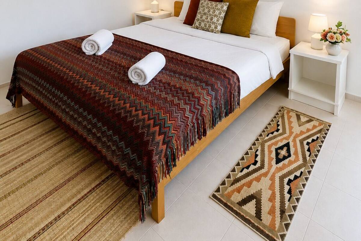 Una cama bien hecha con un edredón estampado, dos mesitas de noche con lámparas, un adorno de pared y una alfombra tejida.