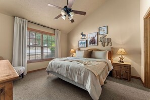 A bedroom with a bed, two bedside tables, a ceiling fan, a window with curtains, and wall-mounted pictures.
