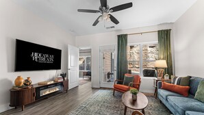 A modern living room with a flat-screen TV, a ceiling fan, a fireplace, a sofa, a coffee table, and a view of a stone wall through the window. - Pool Steps Away! Perfect for Families! Walk-in! (Branson)