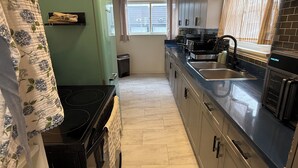 A narrow kitchen with a green refrigerator, a black stove, a sink, and a window with curtains. - Willow & Wildflower Retreat | Cozy, Unique, Family Stay (Edmonton)