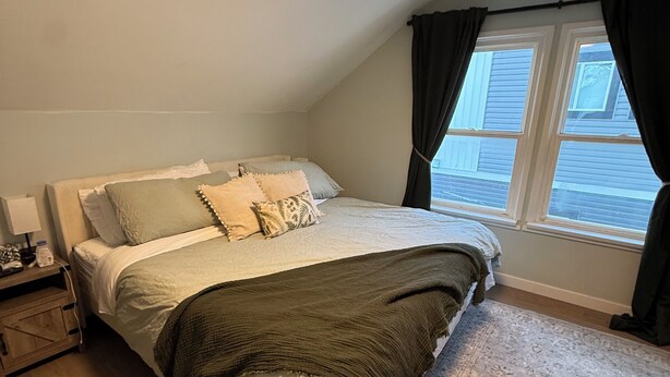 A bedroom with a bed, a nightstand, a window with curtains, and a view of the outside. - Willow & Wildflower Retreat | Cozy, Unique, Family Stay (Edmonton)