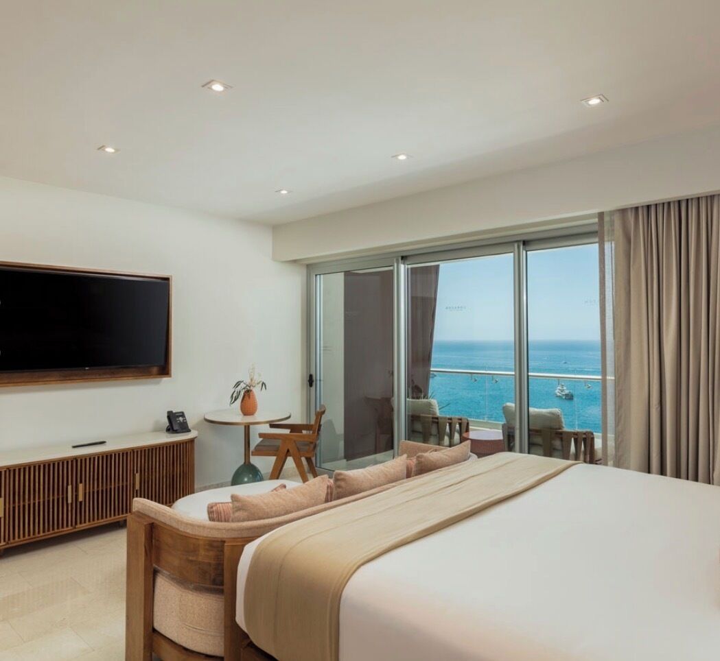 A hotel room with a large bed, a TV, a balcony with a view of the sea, and a small table with chairs.