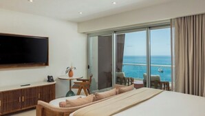 Quarto de hotel com uma cama grande, televisão, varanda vista para o mar e uma pequena mesa com cadeiras.