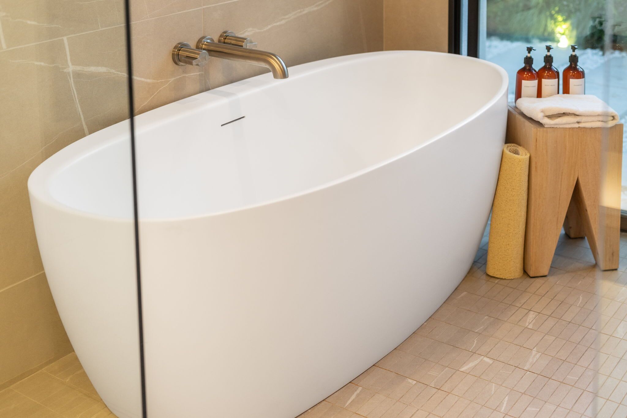 A modern bathroom with a freestanding bathtub, a wooden stool with toiletries, and a glass partition.