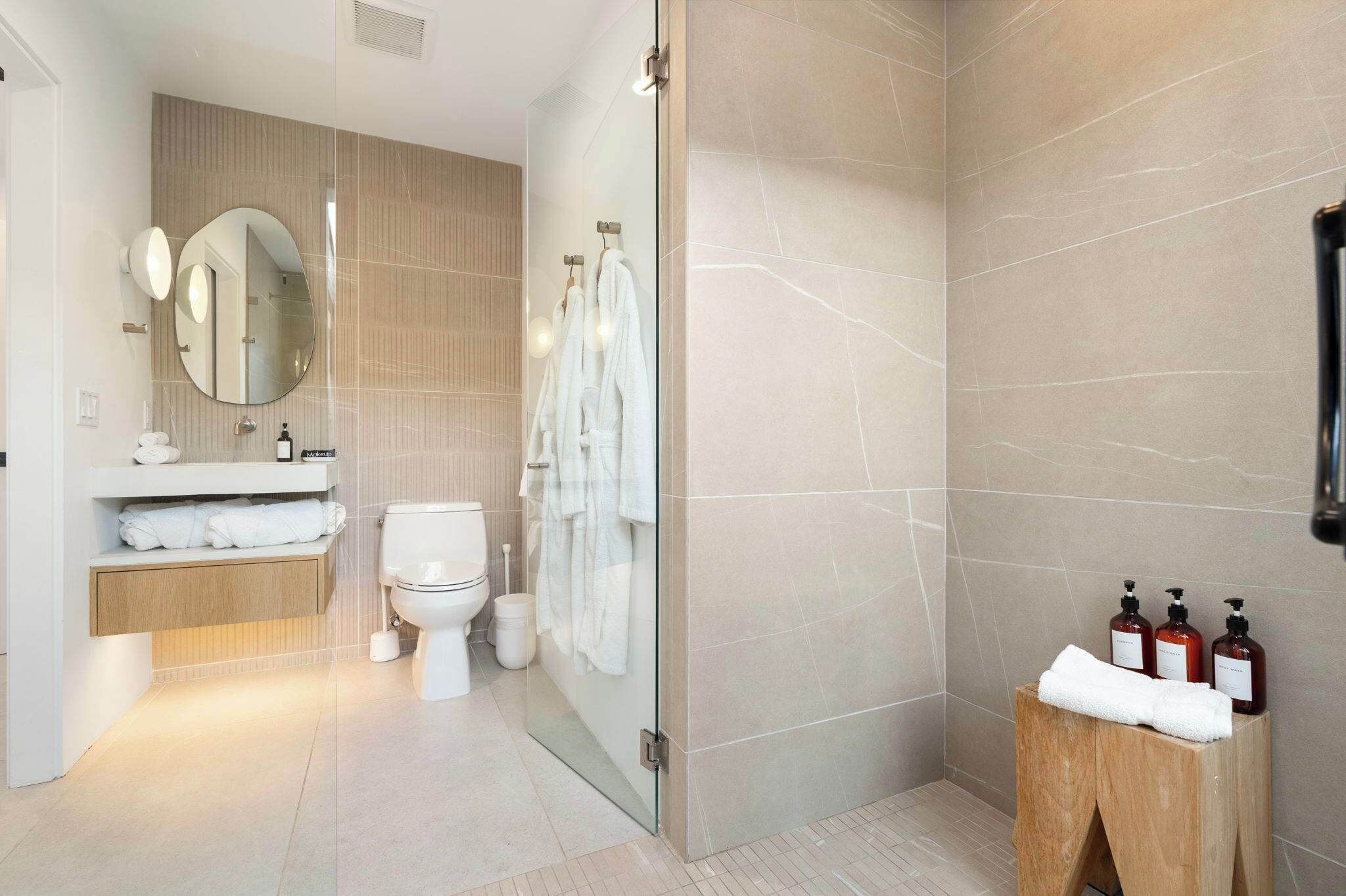 A modern bathroom with a glass shower enclosure, a white toilet, a vanity with a mirror, and a wooden stool with toiletries.