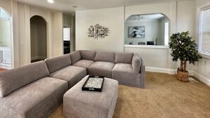 A living room with a sectional sofa, a small ottoman, a decorative wall piece, and a potted plant. - Modern and Stylish Retreat with Game Room (Tigard)