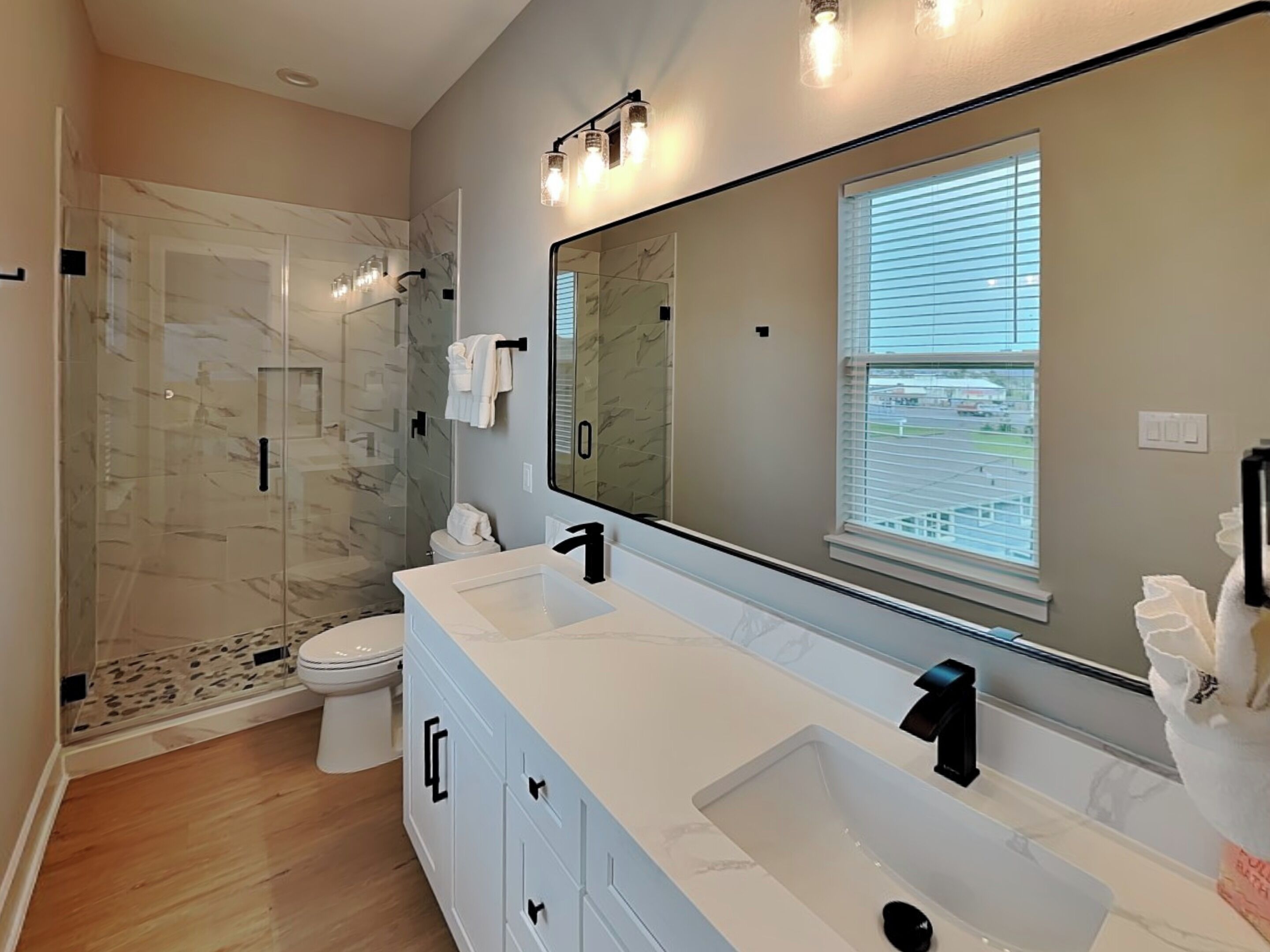 Un baño moderno con un espejo grande, dos lavamanos, cabina de ducha y ventana con persianas.