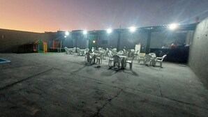 Un espace extérieur aménagé avec des chaises et des tables en plastique, une petite structure de jeux et un terrain de basket-ball.