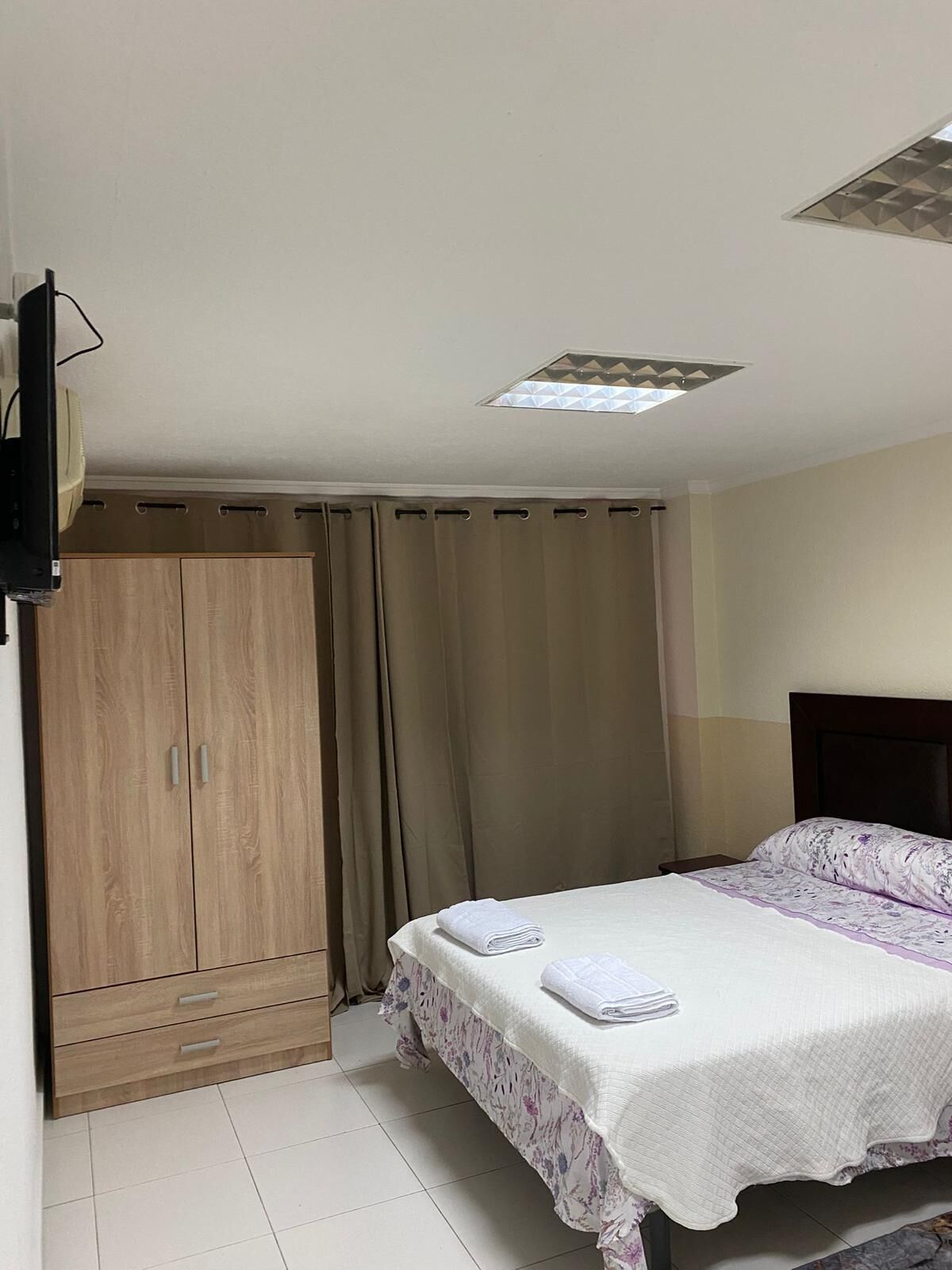 Une chambre à coucher avec un lit, une armoire, une télévision et une fenêtre avec des rideaux.
