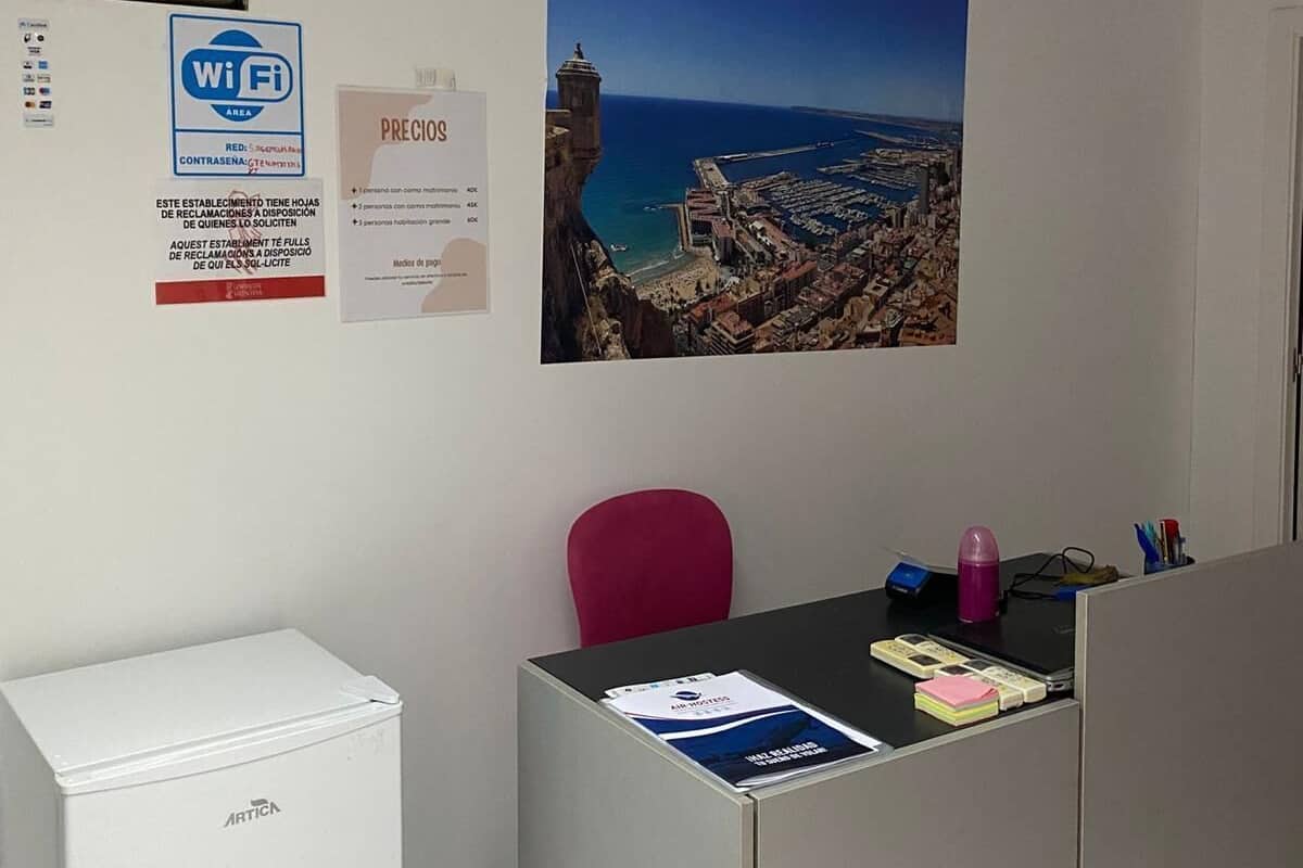 A room with a desk, a red chair, a white air conditioner, a Wi-Fi sign, a price sign, and a scenic coastal photograph on the wall.
