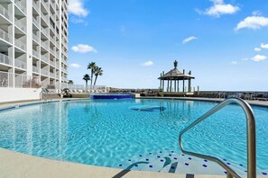 A swimming pool with a metal ladder, lounge chairs, palm trees, and a gazebo. - Beachfront Corner Unit Condo | Pool | Sleeps 6 (Orange Beach)